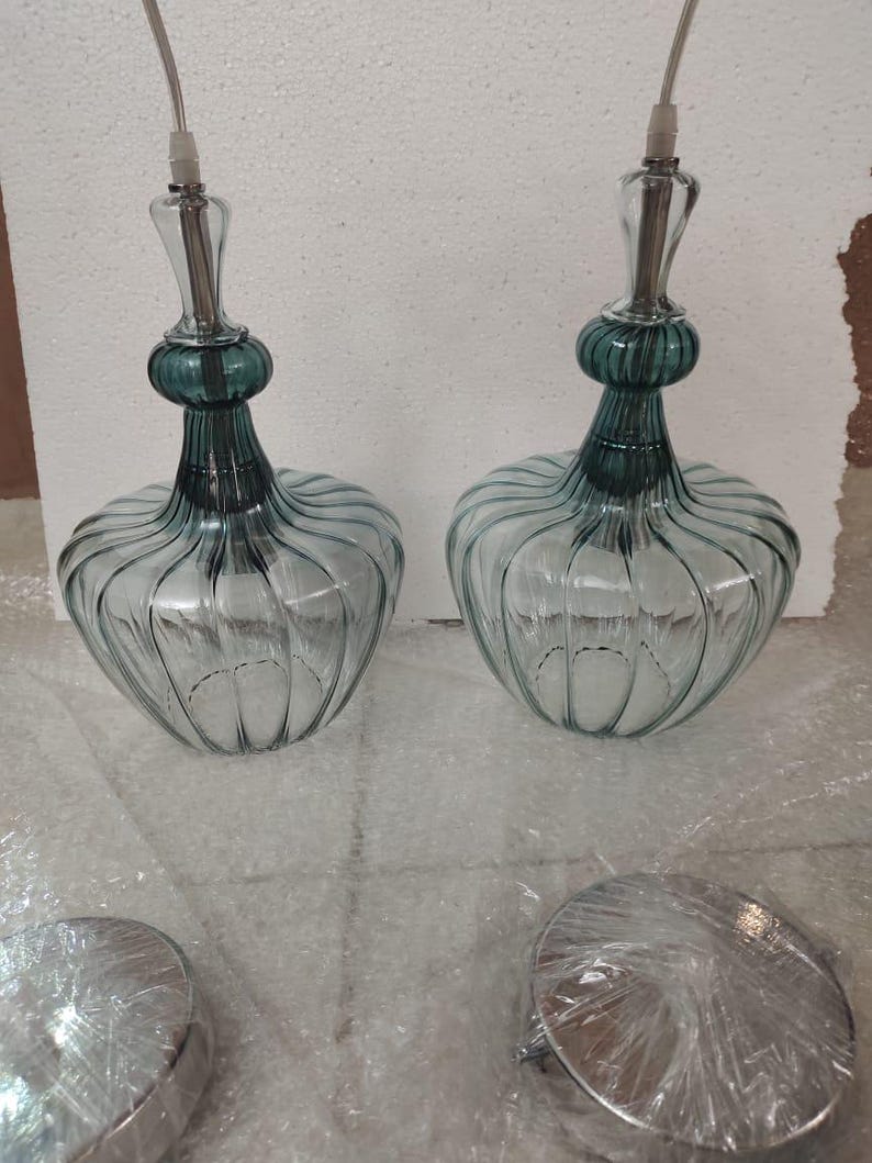 May include: Two glass pendant lights with a teal green top and a clear, ribbed glass body. The lights have a decorative shape and are suspended by a silver cord. Two silver ceiling mounts are in the foreground.