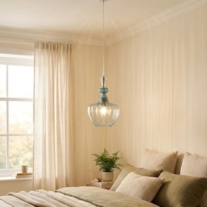 May include: A clear glass pendant light with a ribbed shade and teal accents hangs from the ceiling. The light fixture is suspended above a bed with pillows and a green comforter. The room has beige walls and a window with sheer curtains.