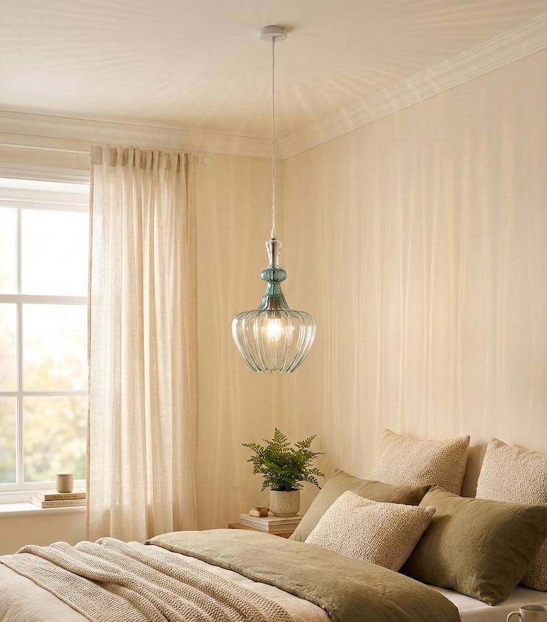 May include: A clear glass pendant light with a ribbed shade and teal accents hangs from the ceiling. The light fixture is suspended above a bed with pillows and a green comforter. The room has beige walls and a window with sheer curtains.