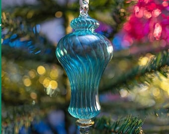 Puede incluir: Un adorno navideño de cristal color turquesa con un diseño en espiral y detalles dorados. El adorno cuelga de una cinta blanca sobre un fondo borroso de un árbol de Navidad con luces de colores.