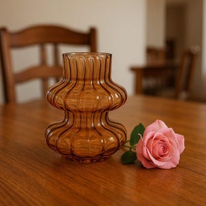 May include: Amber-colored glass vase with a unique, layered design. The vase is sitting on a wooden table next to a pink rose. The vase has a ribbed texture and a rounded shape, perfect for home decor.