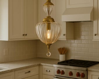 May include: A clear glass pendant light with a ribbed, rounded shade and brass-colored metal accents hangs in a kitchen. The light fixture has a teardrop-shaped crystal at the bottom and a visible lightbulb. The kitchen features white cabinets and a stainless steel stove.
