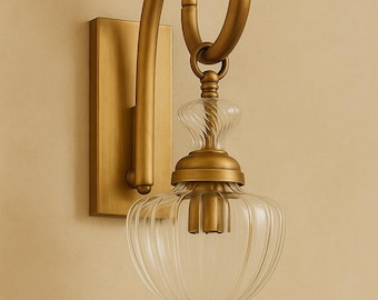 May include: A wall sconce featuring a brushed brass finish rectangular backplate and a curved arm that supports a clear, ribbed glass shade. The fixture includes a decorative brass finial at the bottom and a matching brass neck connecting the glass.