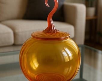 May include: A decorative glass jar with a round body and a lid. The jar is a warm amber color with a swirl pattern. It has a curved, orange handle on top. The jar sits on a glass table, reflecting the light.