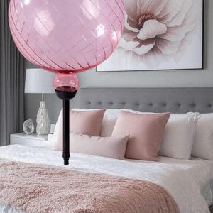 May include: Pink glass globe pendant light with a black stem, hanging above a bed with pink and white bedding. The light has a crisscross pattern. A large floral print hangs on the wall.
