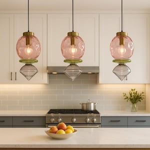 May include: Three pendant lights with pink glass globes, brass accents, and geometric grey and green glass elements. The lights hang in a kitchen setting with white cabinets and a stainless steel range.