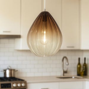 May include: A teardrop-shaped pendant light with a ribbed, ombre glass shade. The shade transitions from a dark brown at the top to a clear, light color at the bottom. The light fixture hangs from a white cord in a kitchen setting.