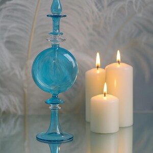 May include: A decorative blue glass bottle with a teardrop-shaped stopper and a round body, next to three lit white pillar candles. The bottle and candles are reflected on a mirrored surface.