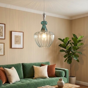May include: A clear glass pendant light with a ribbed, bell-shaped shade and teal accents. The light hangs from a silver-colored fixture. The light is suspended above a green velvet sofa with decorative pillows.