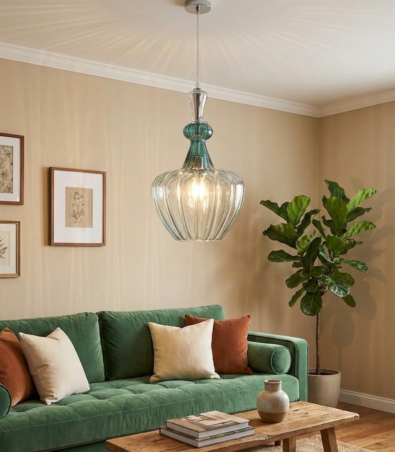 May include: A clear glass pendant light with a ribbed, bell-shaped shade and teal accents. The light hangs from a silver-colored fixture. The light is suspended above a green velvet sofa with decorative pillows.