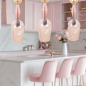 Pode incluir: Três luminárias pendentes com um esquema de cores rosa e dourado pendem sobre uma ilha de cozinha. Cada luminária apresenta uma sombra de vidro transparente em forma de gota, acentuada por elementos decorativos rosa e dourado. A cozinha tem armários rosa e bancos rosa.