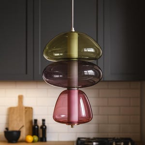 May include: A contemporary pendant light with three coloured glass shades. The top shade is olive green, the middle is purple, and the bottom is pink. The light hangs from a white cord, suitable for a kitchen or dining room. The shades are made of glass.