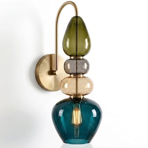 May include: A wall sconce featuring a brushed brass finish metal arm and circular wall plate. The fixture includes four stacked glass elements in olive green, grey, amber, and teal, with a single exposed filament bulb inside the teal base.