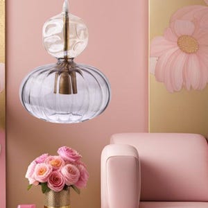 Puede incluir: Una lámpara colgante de cristal transparente con una base redondeada y acanalada y una parte superior bulbosa. La lámpara tiene un acento de metal color latón y cuelga en una habitación con paredes rosas y un sofá rosa. Un ramo de rosas rosas está en un jarrón dorado.