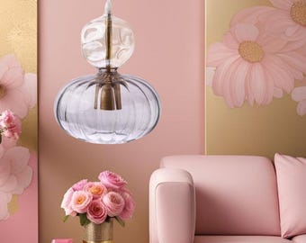 Puede incluir: Una lámpara colgante de cristal transparente con una base redondeada y acanalada y una parte superior bulbosa. La lámpara tiene un acento de metal color latón y cuelga en una habitación con paredes rosas y un sofá rosa. Un ramo de rosas rosas está en un jarrón dorado.