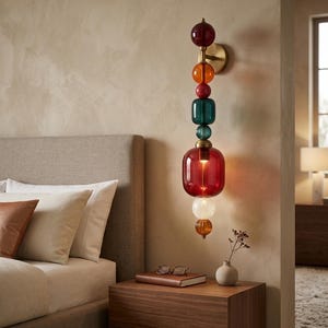 May include: A multi-colored glass wall sconce with a brass fixture. The sconce features a series of glass orbs and rectangular shapes in red, orange, green, and clear colors. The lamp is mounted on a beige wall next to a wooden nightstand.