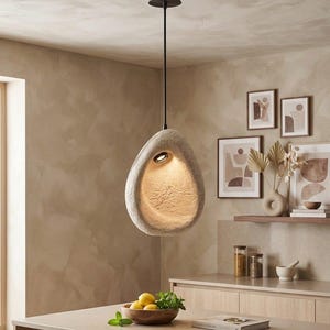 May include: A pendant light fixture featuring a textured, off-white, stone-like organic shape with a hollow center. A single light source is recessed within the interior, casting a warm glow downward. It hangs from a thin, dark metal rod.