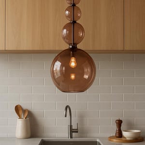 Könnte beinhalten: Eine moderne Pendelleuchte mit einer Reihe brauner Glaskugeln, die an einer schwarzen Stange aufgehängt sind. Die Leuchte ist über einem Küchenbecken installiert, mit einer weißen Fliesenspiegel und Holzschränken im Hintergrund.