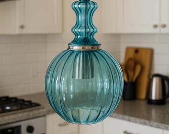 May include: A teal glass pendant light with a ribbed, spherical shade and a decorative top. The light fixture hangs from a black cord and is set against a white kitchen backdrop. The light fixture is a decorative element for a kitchen.
