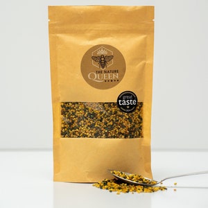May include: A brown paper bag containing bee pollen, with a clear window to view the contents. The bag features a circular label with a bee and the text "THE NATURE QUEEN". A spoon holds some pollen, with a small amount spilled. A "great taste" sticker is also visible.