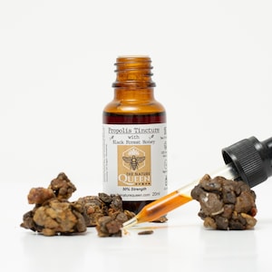 May include: A brown glass bottle with a dropper filled with a dark liquid. The bottle label reads "Propolis Tincture with Black Forest Honey" and features a bee illustration. The label also includes the text "The Nature Queen" and "50% Strength".  There are several brown, irregularly shaped pieces of propolis resin in the image.