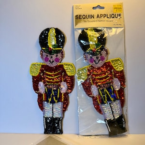 May include: Two red and gold sequined Christmas toy soldier appliques. The appliques are on a white background with a price tag that reads "Sequin Applique An Excellent Fashion Accent 1 Pc. Per Pkg."