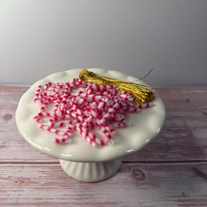 May include: A white ceramic cake stand holds a collection of red and white striped beads and a gold tassel. The beads are scattered across the stand, creating a festive and decorative display. The stand is on a light wood surface.