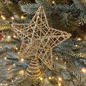 May include: A gold glitter star tree topper with a spiral base. The star is made of a web-like design and is placed on a green Christmas tree with small white lights.