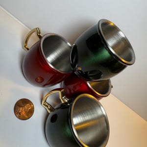 May include: Four metal mugs with gold handles. Two mugs are red and two are green. The mugs are stacked on top of each other.