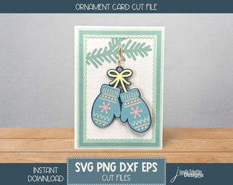 May include: A holiday-themed card featuring a pair of light blue mittens with pink snowflake and zigzag patterns. The mittens are suspended from a green pine branch and a yellow bow. The card has a white background with a green border. The text "ORNAMENT CARD CUT FILE" is at the top.