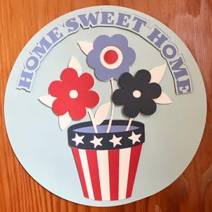 Puede incluir: Un letrero redondo azul claro con las palabras "HOME SWEET HOME" en letras azules. El letrero presenta un jarrón con rayas rojas y blancas, que contiene tres flores en rojo, azul y negro. Hay hojas blancas detrás de las flores.