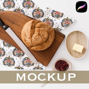 May include: A round loaf of bread sits on a wooden cutting board next to a bread knife. A patterned floral napkin and small dishes of butter and jam complete the breakfast scene. The word "MOCKUP" is displayed in large, bold letters.