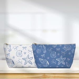 May include: Two zippered cosmetic bags with a nautical theme. One bag is white with blue seashell, starfish, and seahorse patterns. The other bag is blue with white seashell, starfish, and seahorse patterns. Both bags have the name "Taylor" printed on them.
