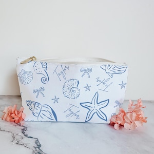 May include: White canvas cosmetic bag with a blue seashell, seahorse, starfish, and bow pattern. The name "Taylor" is written in a script font. The bag has a gold zipper and is decorated with pink flowers.