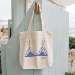 Puede incluir: Bolso tote de color crema con un diseño de sujetador azul. El diseño del sujetador presenta delicados detalles de encaje y el nombre "Taylor" impreso en una fuente cursiva. El bolso está colgado de una estructura de madera azul claro.