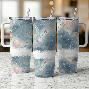 May include: Three stainless steel travel mugs with handles and straws. The mugs feature a celestial design with watercolor-style clouds, stars, and planets in shades of blue, pink, and gold. The bottom half of each mug is white.