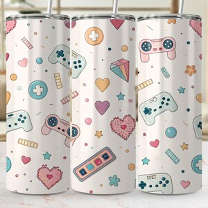 May include: A cylindrical tumbler with a white background featuring a repeating pattern of pastel-colored video game controllers, hearts, diamonds, stars, and circular icons. The design includes soft pink, blue, and orange accents throughout.