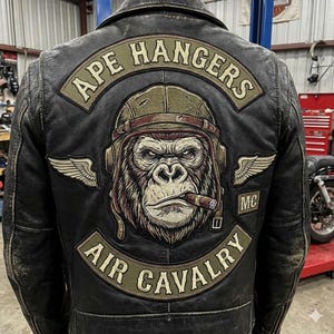 Può includere: Giacca in pelle nera con toppe. La schiena presenta una toppa grande con un gorilla con elmetto e sigaro, con le scritte "APE HANGERS" e "AIR CAVALRY" in testo arcuato. Toppe con ali su entrambi i lati.