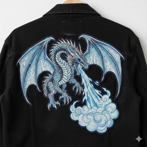 May include: Black denim jacket featuring a large embroidered patch of a blue dragon breathing ice. The dragon has detailed wings and is set against a cloud of ice. The jacket is a classic style with a button closure.
