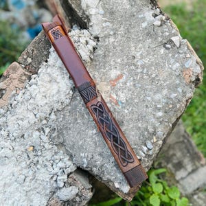 May include: A brown leather knife sheath with intricate Celtic knotwork carvings. The sheath is a rich, dark brown color and features a decorative design. The sheath is designed to hold a knife and is made of durable leather.