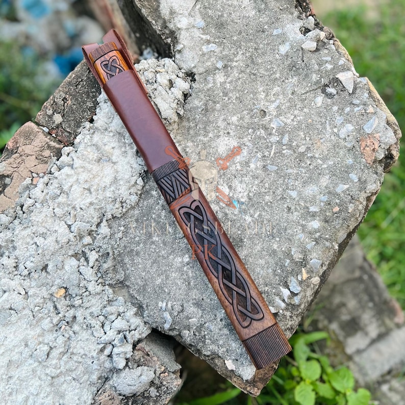 May include: A brown leather knife sheath with intricate Celtic knotwork carvings. The sheath is a rich, dark brown color and features a decorative design. The sheath is designed to hold a knife and is made of durable leather.