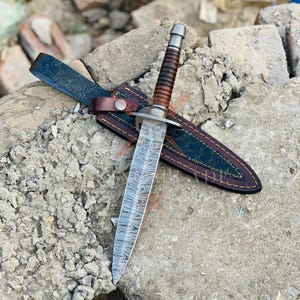 Puede incluir: Una daga de acero de Damasco con una hoja estampada, un mango envuelto en cuero marrón y una guarda de metal plateado. La daga descansa en una funda de cuero negro con costuras marrones y una trabilla de cuero.