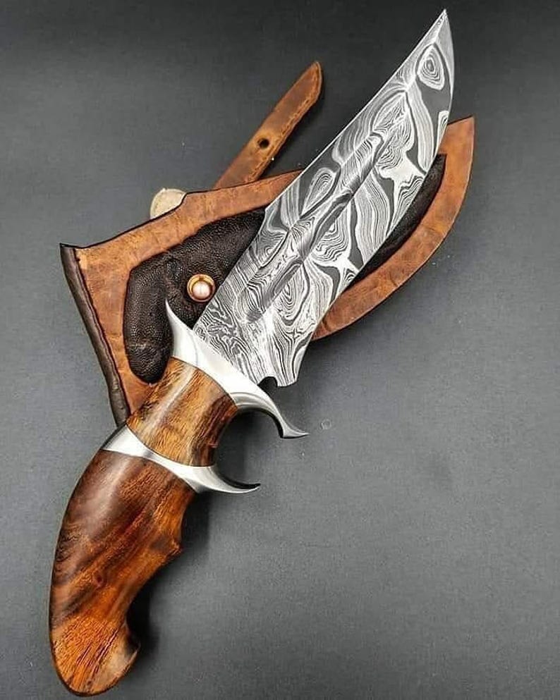 May include: A Damascus steel hunting knife with a wooden handle and a leather sheath. The blade has a unique pattern, and the handle is a rich brown color. The knife features a silver bolster and guard. The sheath is brown leather.