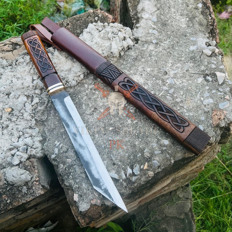 May include: A handcrafted knife with a wooden handle and a stainless steel blade, accompanied by a leather sheath. The handle features an intricate Celtic knot design. The sheath is brown with a carved Celtic knot pattern. The knife is approximately 12 inches long.