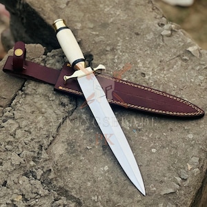 May include: A dagger with a polished steel blade and a cream-colored handle, accented with gold-colored metal. The dagger rests on a brown leather sheath with a stitched border. The image is set against a textured stone surface.