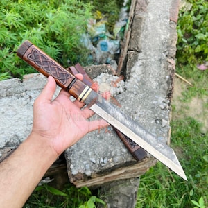 May include: A handcrafted knife with a wooden handle featuring an intricate Celtic knot design. The blade is made of steel. The knife is displayed on a concrete surface with greenery in the background. The text "VIKINGSBLADE" is visible.