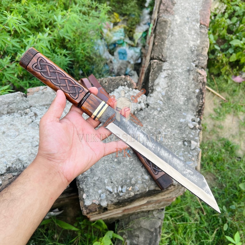 May include: A handcrafted knife with a wooden handle featuring an intricate Celtic knot design. The blade is made of steel. The knife is displayed on a concrete surface with greenery in the background. The text "VIKINGSBLADE" is visible.