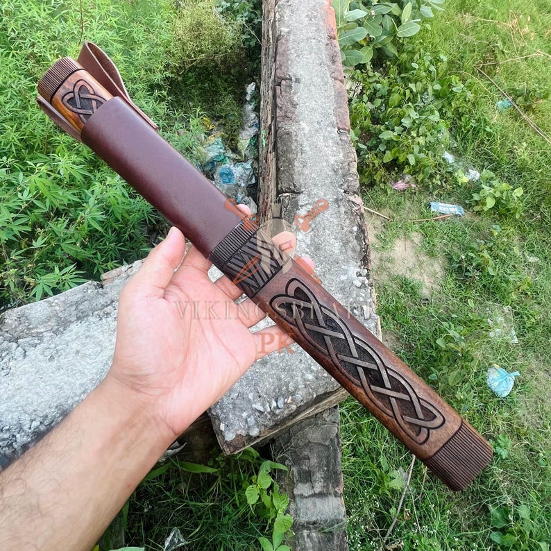 May include: A brown leather sheath with intricate Celtic knotwork and a matching handle. The sheath is designed to hold a sword or knife. The handle and sheath are made of wood and leather. The image shows the product in a natural outdoor setting.