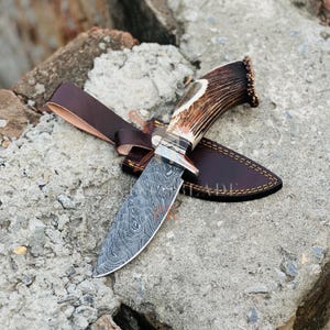 Peut inclure: Un couteau de chasse en acier de Damas avec un manche en corne marron et un étui en cuir. La lame présente un motif tourbillonnant unique. L'étui est marron avec une bordure cousue et une lanière en cuir.