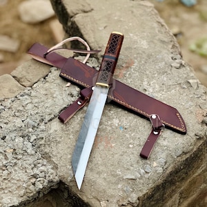 Puede incluir: Un cuchillo artesanal con una hoja de acero inoxidable y un mango decorativo con un diseño de nudo celta. El cuchillo va acompañado de una funda de cuero marrón con trabilla para cinturón. La hoja mide aproximadamente 25 cm.